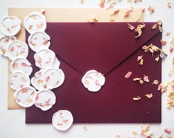 Peut inclure: Une enveloppe bordeaux avec un sceau de cire blanc et une enveloppe beige avec plusieurs sceaux de cire blancs avec des accents de fleurs séchées. L'arrière-plan est blanc avec des pétales de fleurs séchées éparpillés.