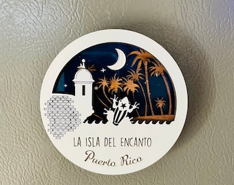 May include: White circular wooden sign with a black and brown design. The design features a crescent moon, palm trees, a building with a tower, and a frog. The text "LA ISLA DEL ENCANTO Puerto Rico" is written in brown on the sign.