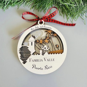 May include: White round wooden ornament with a laser cut design of palm trees, a fort, and a coqui frog. The ornament has the text "La Isla del Encanto" at the top and "Familia Valle Puerto Rico" at the bottom.