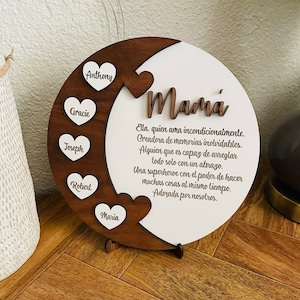 May include: A wooden circle with a white background and a brown crescent moon shape. The crescent moon has five white hearts with names written on them: Anthony, Gracie, Joseph, Robert, and Maria. The white background has the word "Mamá" written in cursive and a Spanish message about a mother's love.