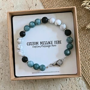 May include: A beaded bracelet featuring alternating white marble, dark green, and black porous lava stone beads with silver-toned spacers and a lobster claw clasp, presented on a white display card with the text CUSTOM MESSAGE HERE.