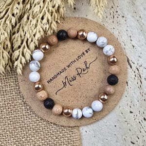 May include: A bracelet made with white marble, wood, lava rock, and rose gold beads. The bracelet is on a brown coaster with the text "Handmade with love by Miss Roh".