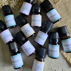 May include: A collection of 11 small glass bottles with black caps, each containing a different essential oil. The bottles are labelled with the names of the oils, including Eucalyptus, Rose Geranium, Sweet Orange, Frankincense, Lavender, Peppermint, and more. The bottles are arranged on a light brown surface.