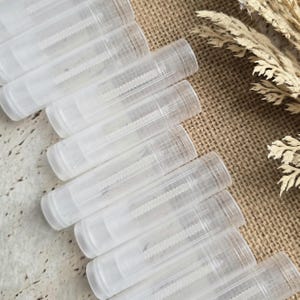May include: A collection of empty, translucent white plastic lip balm tubes arranged in a diagonal row. Each cylindrical container features a removable cap and a visible internal twist mechanism with a ridged white plastic stem for product dispensing.