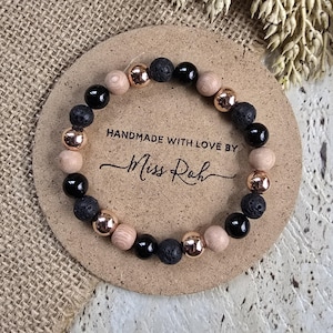 May include: A circular beaded bracelet featuring alternating black lava stone beads, smooth black onyx beads, light tan wooden beads, and metallic rose gold-toned beads. The bracelet is displayed on a round tan card with the text HANDMADE WITH LOVE BY Miss Roh.