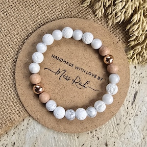 May include: A beaded bracelet featuring white porous lava stone beads, smooth white marble-like beads, light brown wooden beads, and two metallic rose gold-coloured spherical beads. The bracelet is displayed on a round card with the text HANDMADE WITH LOVE BY Miss Roh.