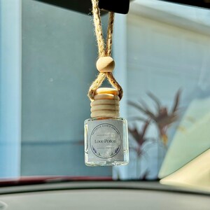 Op de afbeelding: Een heldere glazen auto luchtverfrisser met een houten dop en een natuurlijke vezel koord om op te hangen. Het label leest "Love Potion" met een lavendel ontwerp. De luchtverfrisser hangt voor een autoruit.