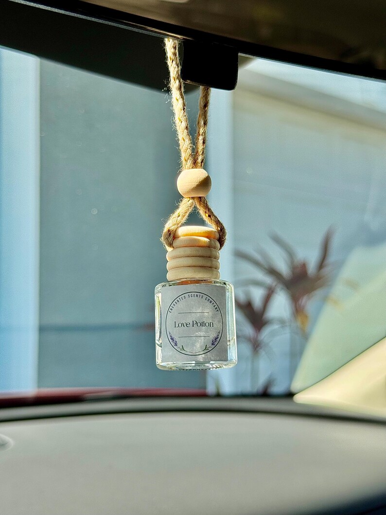 Op de afbeelding: Een heldere glazen auto luchtverfrisser met een houten dop en een natuurlijke vezel koord om op te hangen. Het label leest "Love Potion" met een lavendel ontwerp. De luchtverfrisser hangt voor een autoruit.