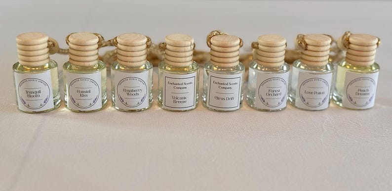 Op de afbeelding: Een verzameling kleine, heldere glazen flesjes met houten doppen en etiketten. Elk flesje bevat een ander parfum, met etiketten met de tekst "Tranquil Bloom", "Eternal Kiss", "Cranberry Woods", "Volcanic Breeze", "Citrus Drift", "Forest Orchard", "Love Potion" en "Peach Dreams".
