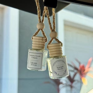 May include: Two small, clear glass bottles with wooden tops and hanging cords. One bottle reads "Volcanic Breeze" and the other "Tranquil Bloom." The bottles are hanging from a car's rearview mirror.
