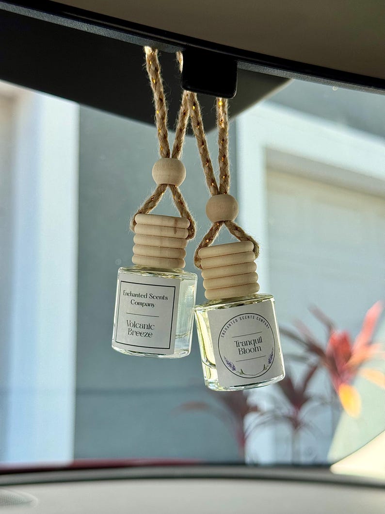 Op de afbeelding: Twee kleine, heldere glazen flesjes met houten doppen en ophangkoorden. Op &eacute;&eacute;n flesje staat "Volcanic Breeze" en op de andere "Tranquil Bloom". De flesjes hangen aan de achteruitkijkspiegel van een auto.