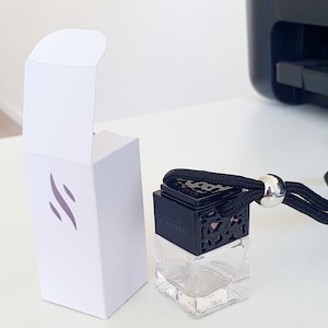 May include: A small, clear glass bottle with a black, decorative lid and a black string attached. The bottle is sitting on a white surface and is in a white box with a logo that says "Scent"