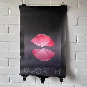 Könnte beinhalten: Ein schwarzes Poster mit einem rosa Blütenblatt-Design und dem Text "KOZO REFLET" in Weiß. Das Poster hängt vor einer weißen Backsteinmauer.