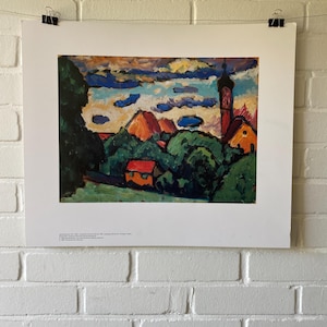 Può includere: Un dipinto di paesaggio incorniciato raffigurante una scena di villaggio con un campanile. L'opera d'arte utilizza pennellate audaci e colori vivaci, tra cui blu, arancione e verde. La stampa è tenuta da clip nere contro un muro di mattoni bianchi.