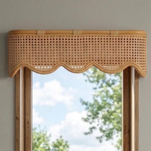 Op de afbeelding: Een lichtbruine houten raamvalance met een geweven rietpatroon en een geschulpte onderrand. De valance is gemonteerd boven een rechthoekig raamkozijn met een natuurlijke houten afwerking, met een kleine vetplant op de vensterbank.