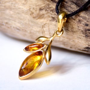 May include: A gold-colored pendant necklace featuring a leaf design with three amber-colored stones. The pendant is suspended from a black cord. The amber stones have a warm, translucent appearance.