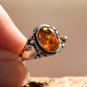 Könnte beinhalten: Ein Silberring mit einem zentralen, ovalen Bernstein in warmem, durchscheinendem Orange. Die Metallfassung weist eine detaillierte, seilartige Struktur sowie kleine kugelförmige Verzierungen entlang der Ränder der Steinfassung auf.