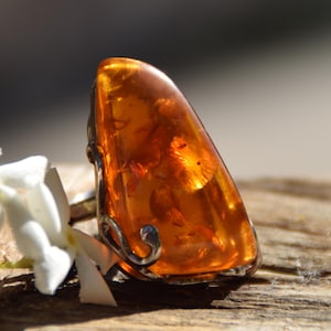 May include: A ring featuring a large, polished, teardrop-shaped piece of translucent orange amber. The amber is set within a silver-toned metal frame that features delicate, curved, wire-like detailing along the side of the stone setting.