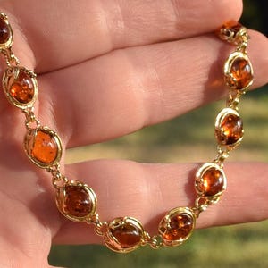 Op de afbeelding: Een goudkleurige schakelarmband met een reeks ovale, doorschijnende barnsteenkleurige stenen. Elke steen is gevat in een decoratieve, getextureerde gouden metalen zetting, verbonden door kleine ronde schakels om een kettingontwerp te vormen.