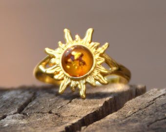 Puede incluir: Un anillo de tono dorado con una piedra preciosa de ámbar circular en el centro, rodeada por un diseño de rayos solares. La banda de metal es lisa y pulida, enmarcando la cálida piedra de color naranja parduzco y translúcido.