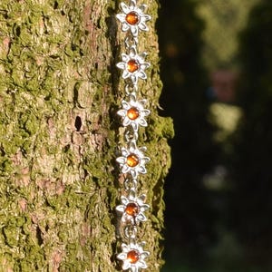 Pode incluir: Uma pulseira de corrente prateada com pingentes em forma de flor com centros de cor âmbar. A pulseira é exibida em um fundo de tronco de árvore texturizado, verde e marrom, com a luz do sol destacando os detalhes.