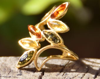 May include: A gold-toned leaf-shaped ring with amber-colored and green-toned stones. The ring has a curved design with multiple leaf-shaped stones in shades of amber and green. The ring is set on a wooden surface.