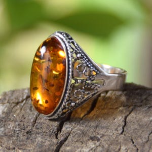 May include: A silver ring featuring a large, oval-shaped, translucent amber gemstone with visible internal inclusions. The metal band displays an intricate, openwork filigree pattern along the sides, supporting the central amber setting.