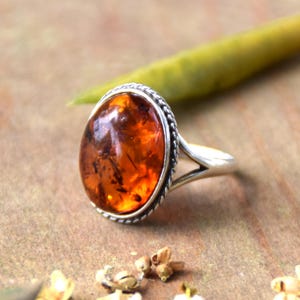 May include: A ring featuring a large, oval-shaped, translucent amber stone with visible internal inclusions. The stone is set in a polished silver bezel with a decorative twisted rope border, mounted on a simple, smooth silver band.