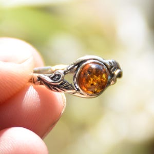 Op de afbeelding: Een zilveren ring met een barnsteenkleurige steen. De ring heeft een bladeren ontwerp op de band en rond de steen. De barnsteenkleurige steen is rond en heeft een warme, gouden tint. De ring wordt in een hand gehouden.