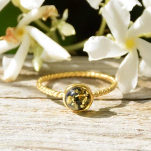 May include: A gold ring with a twisted band and a dark, round stone. The stone is filled with gold flecks. White flowers are in the background, resting on a wooden surface.
