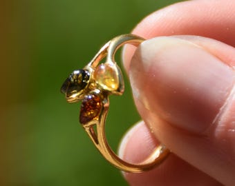 May include: A gold-toned ring featuring three small, teardrop-shaped stones in varying shades of dark green, light yellow, and deep amber. The metal band has a split design that branches out to hold the three distinct, polished gemstone accents.