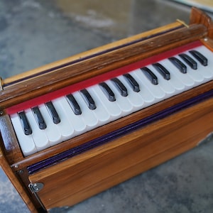 May include: A wooden harmonium with a keyboard of white and black keys. The instrument has a reddish-orange accent above the keys and a purple trim. The wooden case has a natural wood grain finish.