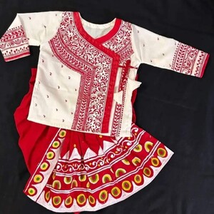 May include: A traditional white and red outfit featuring a long-sleeved top and a skirt. The top has intricate red embroidery around the neckline and sleeves. The skirt has a detailed pattern with red, yellow, and white accents.