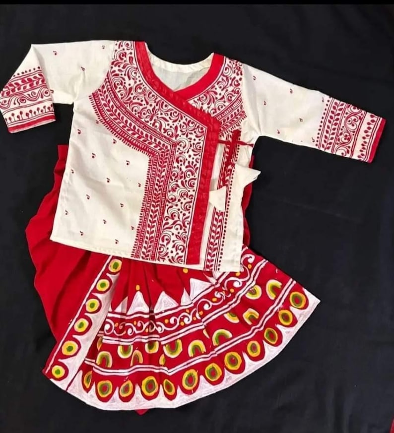May include: A traditional white and red outfit featuring a long-sleeved top and a skirt. The top has intricate red embroidery around the neckline and sleeves. The skirt has a detailed pattern with red, yellow, and white accents.