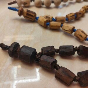 K&ouml;nnte beinhalten: Nahaufnahme von drei Str&auml;ngen Holzperlen. Die Perlen haben verschiedene Formen und Farben, darunter Dunkelbraun, Hellbraun und Off-White. Die Perlen sind auf blauen und braunen Kordeln aufgef&auml;delt. Der Hintergrund ist eine hellfarbige Oberfl&auml;che.