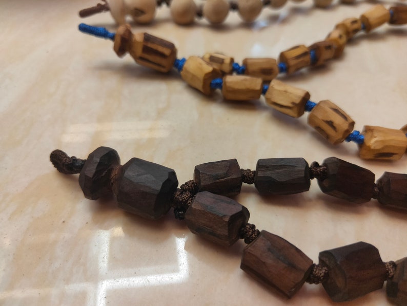 K&ouml;nnte beinhalten: Nahaufnahme von drei Str&auml;ngen Holzperlen. Die Perlen haben verschiedene Formen und Farben, darunter Dunkelbraun, Hellbraun und Off-White. Die Perlen sind auf blauen und braunen Kordeln aufgef&auml;delt. Der Hintergrund ist eine hellfarbige Oberfl&auml;che.