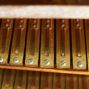 May include: Close-up of a vintage piano's internal mechanism. The image shows a series of rectangular brass-coloured metal bars, each secured with small silver screws. Some bars have stamped text, including the words "FACTORY PREMIER INC."