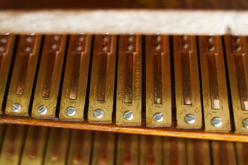 May include: Close-up of a vintage piano's internal mechanism. The image shows a series of rectangular brass-coloured metal bars, each secured with small silver screws. Some bars have stamped text, including the words "FACTORY PREMIER INC."