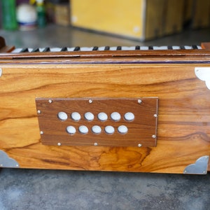 May include: A wooden harmonium with a polished finish. The instrument features a rectangular body with a series of white keys and a dark brown panel with white circular buttons. Silver metal corner accents add detail. The harmonium is a traditional musical instrument.