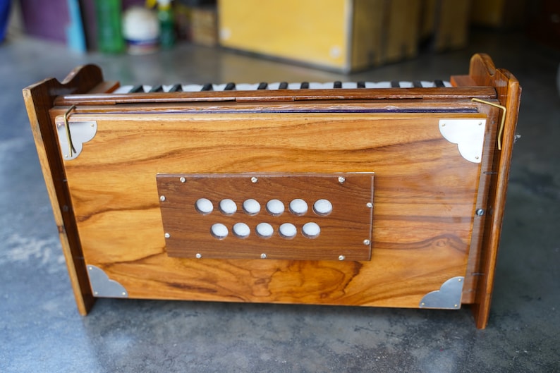 May include: A wooden harmonium with a polished finish. The instrument features a rectangular body with a series of white keys and a dark brown panel with white circular buttons. Silver metal corner accents add detail. The harmonium is a traditional musical instrument.