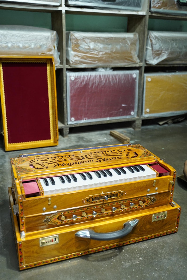 K&ouml;nnte beinhalten: Ein Holzharmonium mit einer schwarz-wei&szlig;en Tastatur, einer roten Filzverkleidung und einem dekorativen geschnitzten Design. Das Harmonium befindet sich in einem braunen Holzkoffer mit einem schwarzen Griff und der Aufschrift "Mayapur Stone" auf der Oberseite.