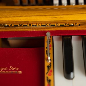 May include: Close-up of a harmonium, a free-reed keyboard instrument. The wooden frame is a warm yellow colour, with black and white keys. The notes are labelled above the keys. The red velvet interior contrasts with the wood. The text "Mayapur Store" is visible.