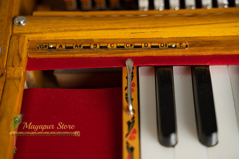 May include: Close-up of a harmonium, a free-reed keyboard instrument. The wooden frame is a warm yellow colour, with black and white keys. The notes are labelled above the keys. The red velvet interior contrasts with the wood. The text "Mayapur Store" is visible.