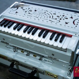 May include: A white harmonium with a decorative carved wooden top and a black keyboard. The harmonium is in a white case with a black handle.