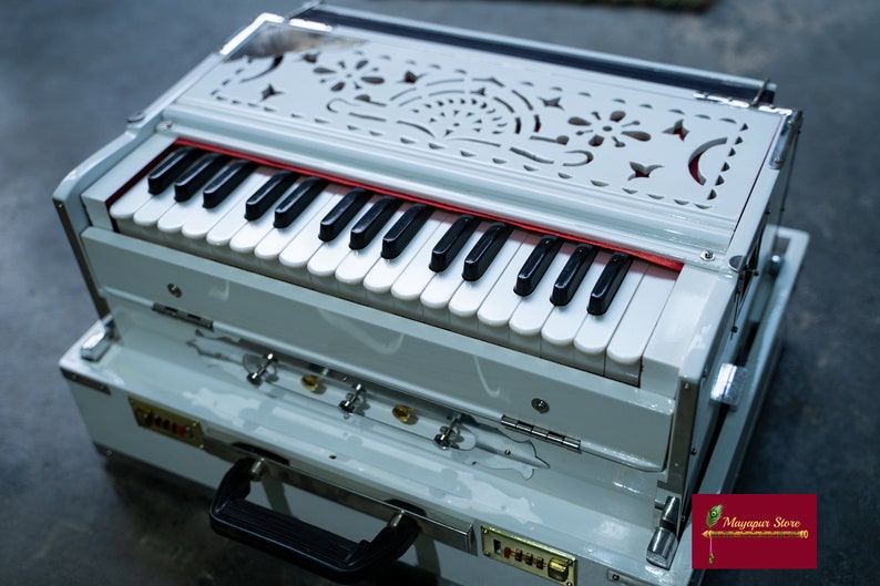 May include: A white harmonium with a decorative carved wooden top and a black keyboard. The harmonium is in a white case with a black handle.