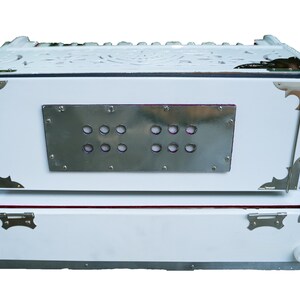 May include: A white harmonium with a silver metal panel featuring a row of round holes. The harmonium has a carved wooden top and silver metal accents.