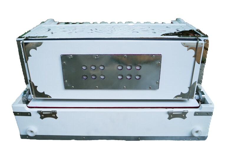 May include: A white harmonium with a silver metal panel featuring a row of round holes. The harmonium has a carved wooden top and silver metal accents.