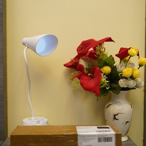 K&ouml;nnte beinhalten: Eine wei&szlig;e Schreibtischlampe mit flexiblem Arm und wei&szlig;em Schirm. Die Lampe steht auf einem braunen Karton mit einem Versandetikett. Eine Vase mit roten und gelben Blumen steht auf einer grauen Oberfl&auml;che.