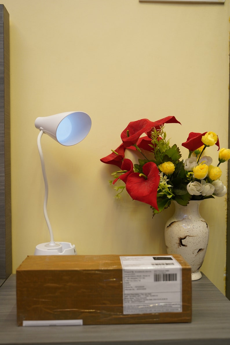 K&ouml;nnte beinhalten: Eine wei&szlig;e Schreibtischlampe mit flexiblem Arm und wei&szlig;em Schirm. Die Lampe steht auf einem braunen Karton mit einem Versandetikett. Eine Vase mit roten und gelben Blumen steht auf einer grauen Oberfl&auml;che.