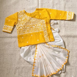 May include: A yellow and white traditional outfit for children. The top is yellow with white floral embroidery and long sleeves. The bottom is white with a yellow patterned border. The outfit is made of a soft fabric.